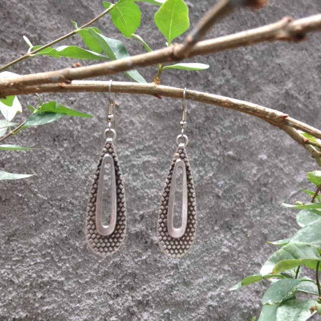 A pair of silver-toned teardrop dangle earrings with textured beaded edges and a central oval cutout, hanging from a tree branch with green leaves against a gray textured wall.