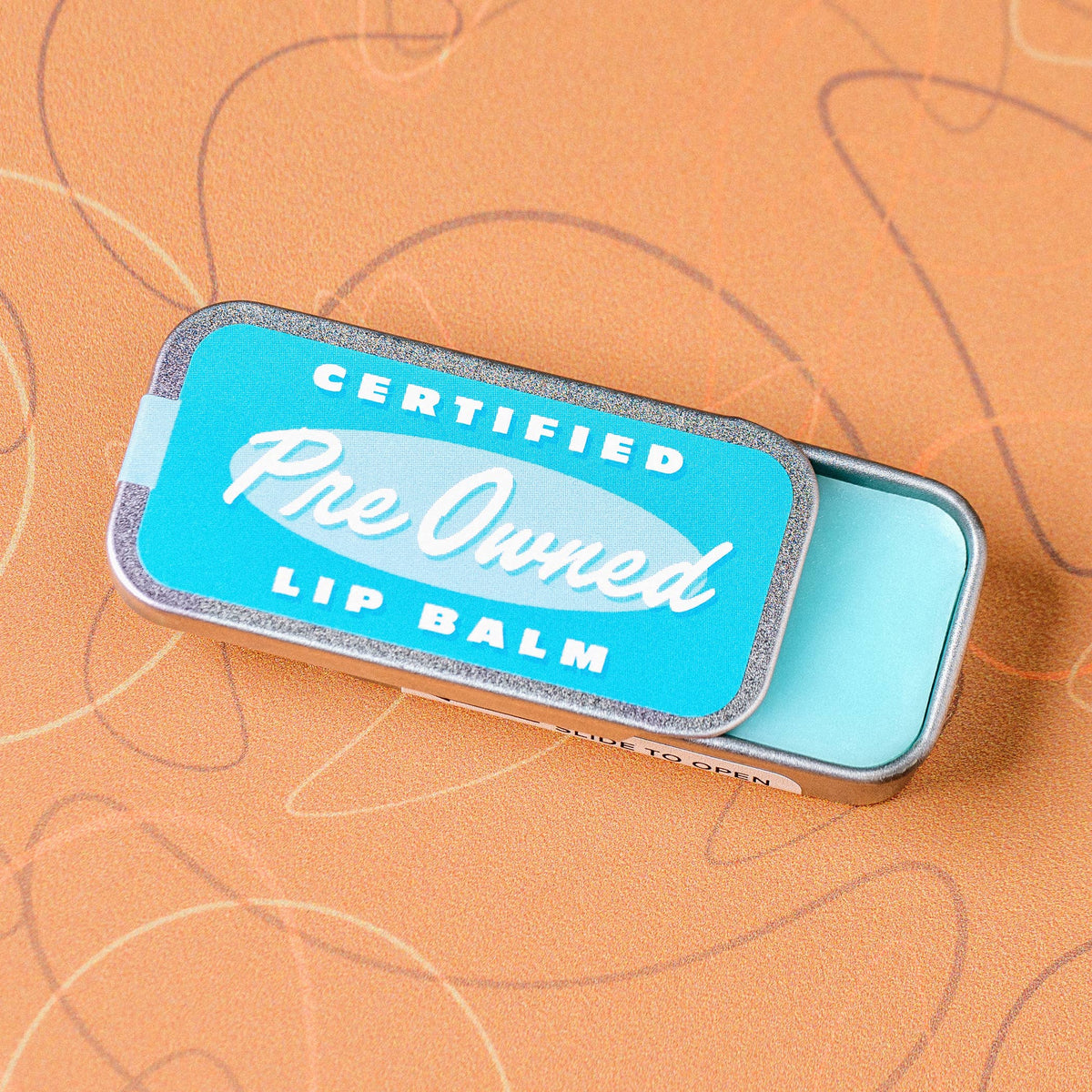 Lip balm tin with 'Certified Pre-Owned' branding on a brown background