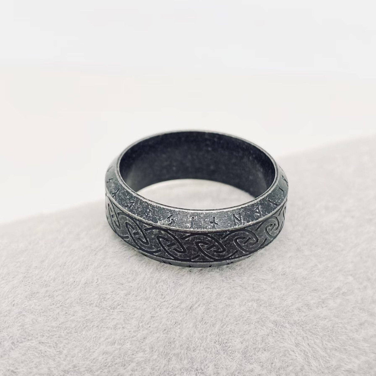 A dark gray metallic ring with an engraved Celtic knot pattern, resting on a textured light gray surface against a white background.