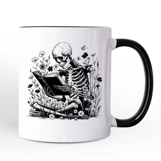 A white mug with a black handle and a graphic of a skeleton reading a book in a patch of flowers, on a white background.
