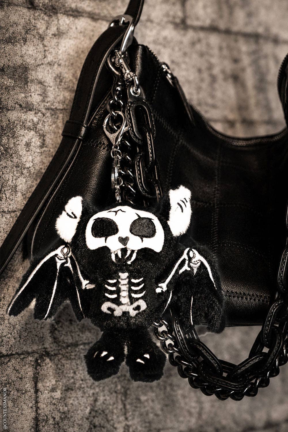 A black skeleton bat plush charm hanging from silver-tone chains, set against a textured gray stone background.
