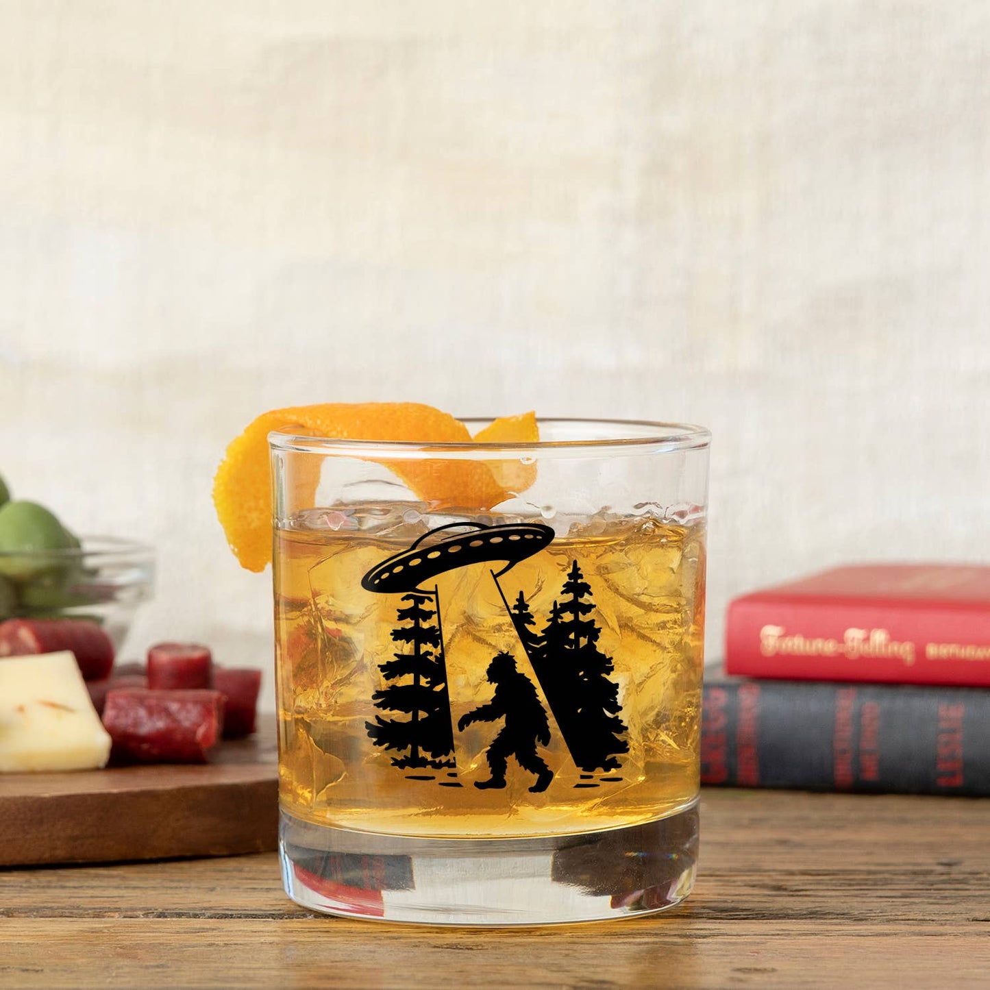 Small glass of beverage with a Bigfoot and UFO design, garnished with an orange peel, on a wooden table.