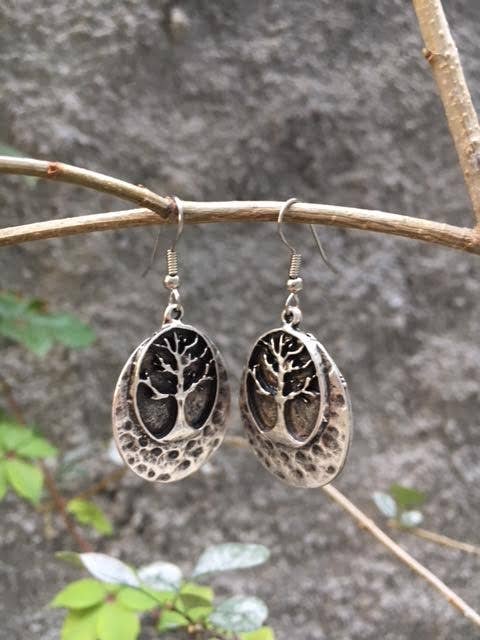 A pair of oval dangling earrings with a tree-of-life motif, hanging from a thin branch against a blurred outdoor background of gray stone and green leaves.