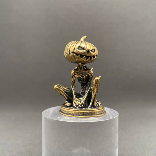 Brass figurine of a skeleton with a pumpkin head on a clear stand against a gray background