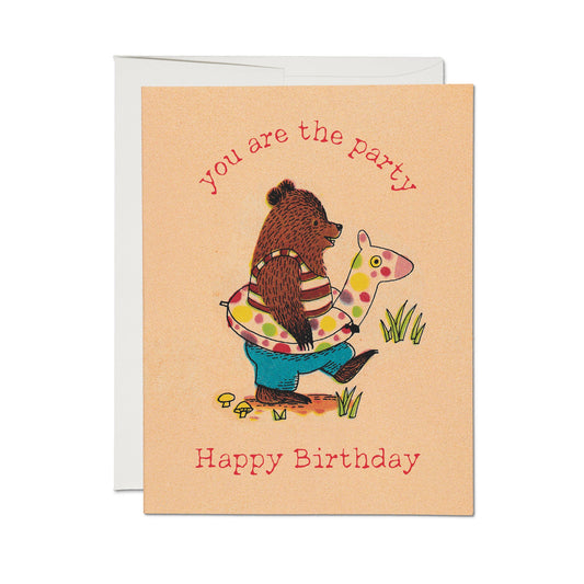 A birthday greeting card with a peach background, featuring a cartoon bear wearing a striped shirt and blue pants while wearing a giraffe raft. The text reads 'you are the party' at the top and 'Happy Birthday' at the bottom.