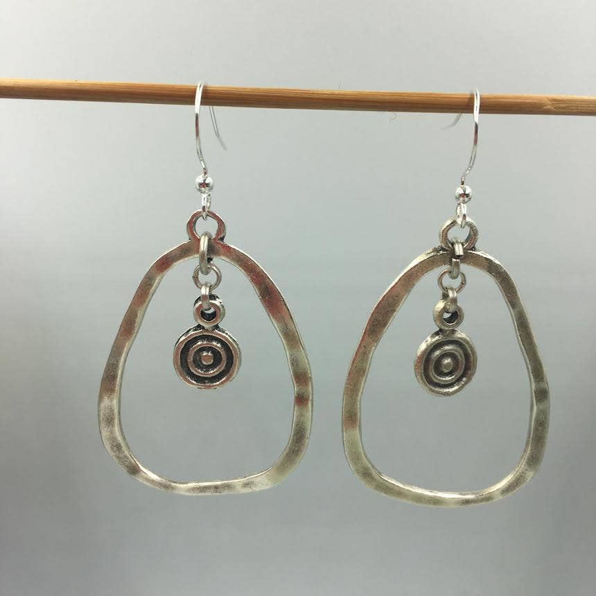 A pair of handmade silver dangle earrings with hammered teardrop-shaped hoops and a small spiral charm, hanging from a wooden dowel against a light gray background.