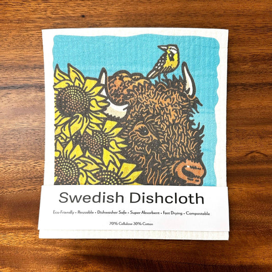 A reusable Swedish dishcloth with a printed illustration of a Highland cow, yellow sunflowers, and a small bird on a turquoise background, laid flat on a wooden surface.