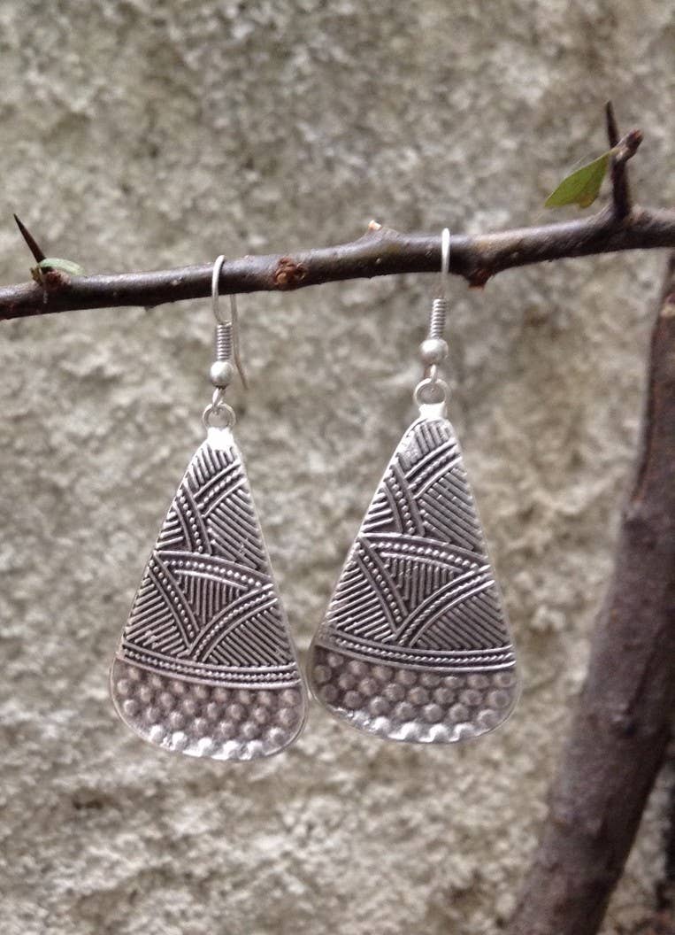 A pair of silver-toned teardrop-shaped dangle earrings with geometric engraved patterns and a textured lower section, hanging from a thin tree branch against a blurred gray stone wall background.