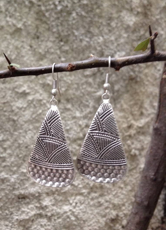 A pair of silver-toned teardrop-shaped dangle earrings with geometric engraved patterns and a textured lower section, hanging from a thin tree branch against a blurred gray stone wall background.