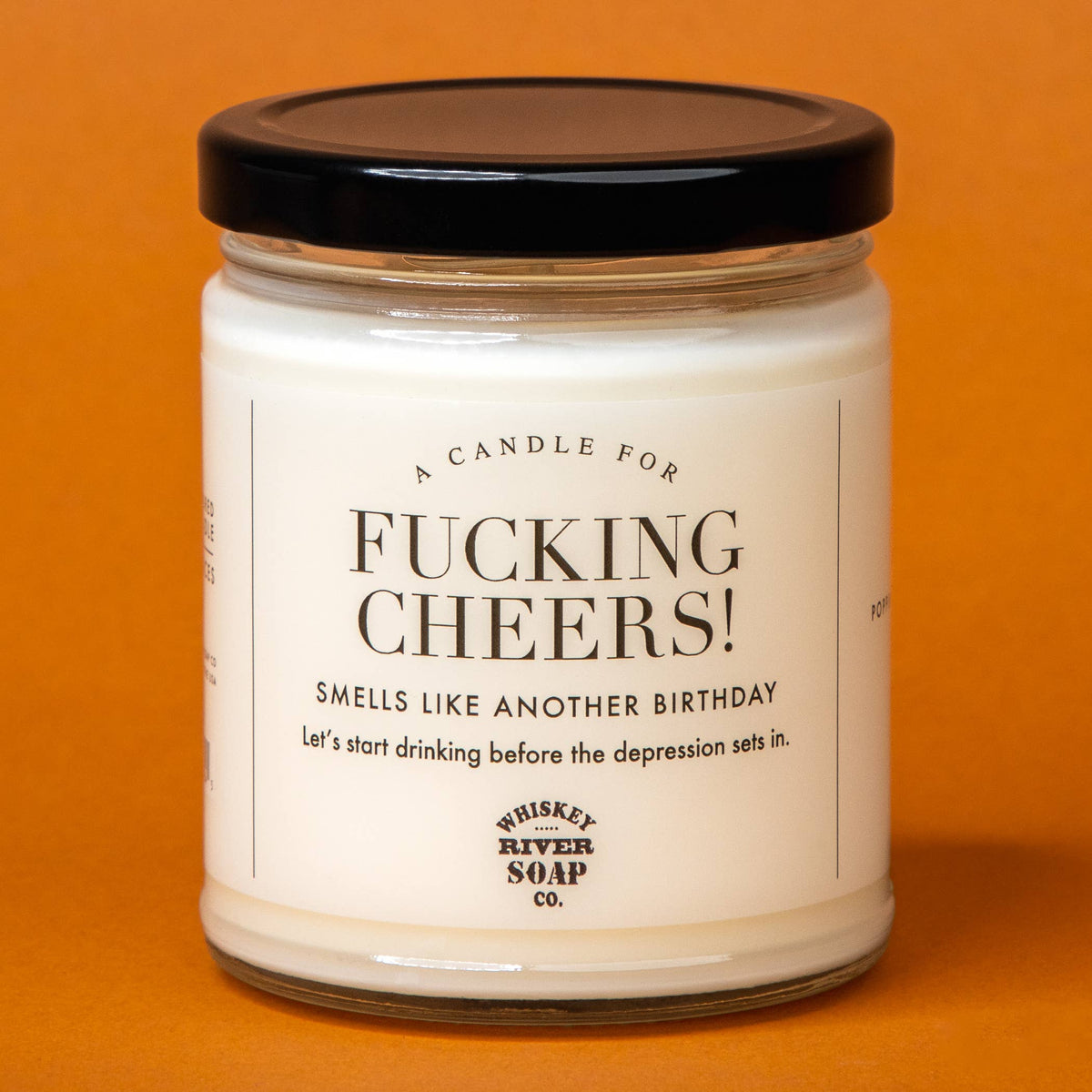 A jar candle with a black lid and a white label reading 'F.CKING CHEERS!' and 'Whiskey River Soap Co.' on a solid orange background.