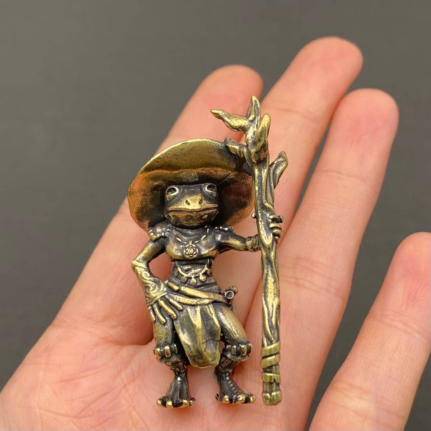 Brass figurine frog with a hat and staff held in a hand against a dark background