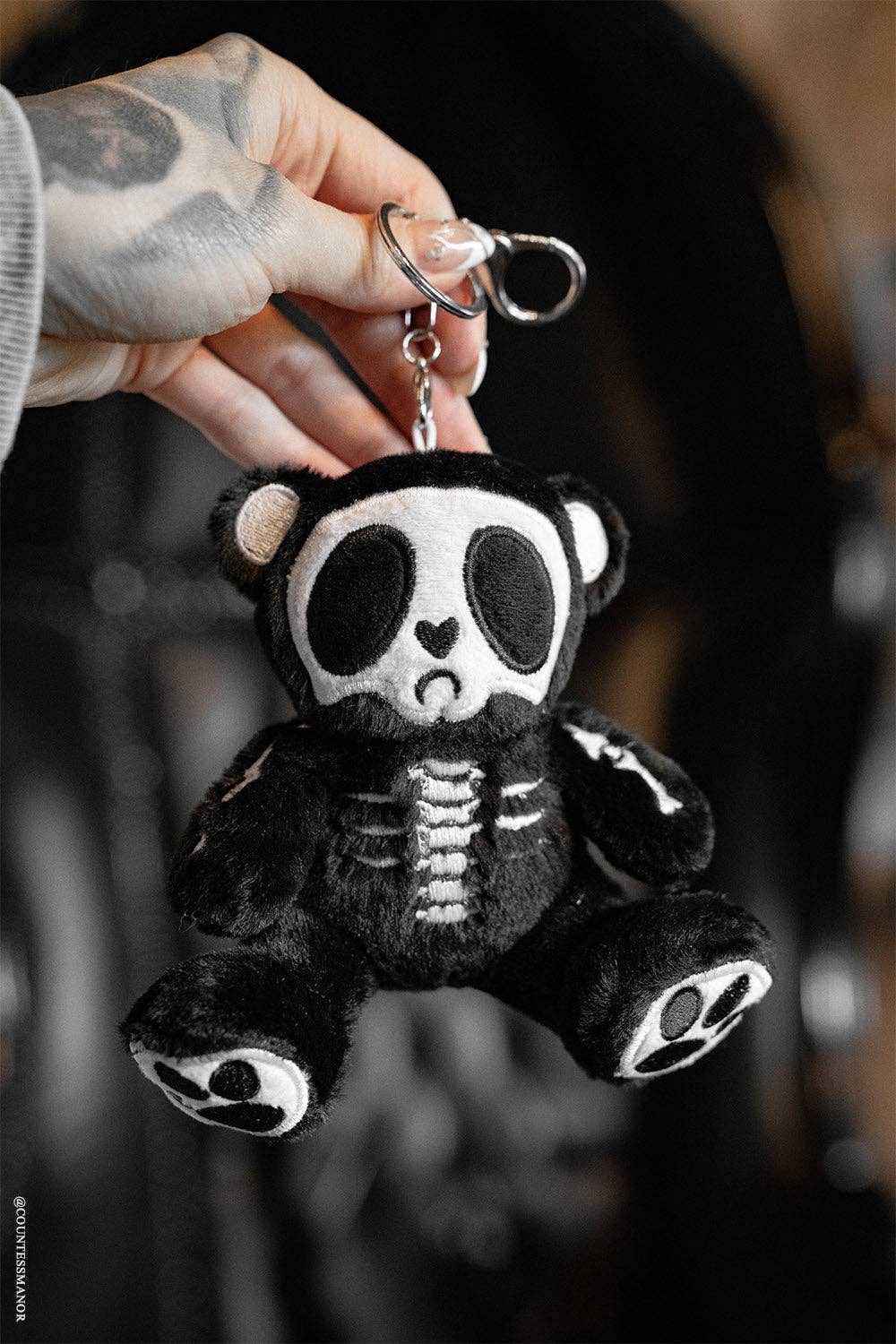 Black and white skeleton teddy bear keychain held by a tattooed hand against a blurred background