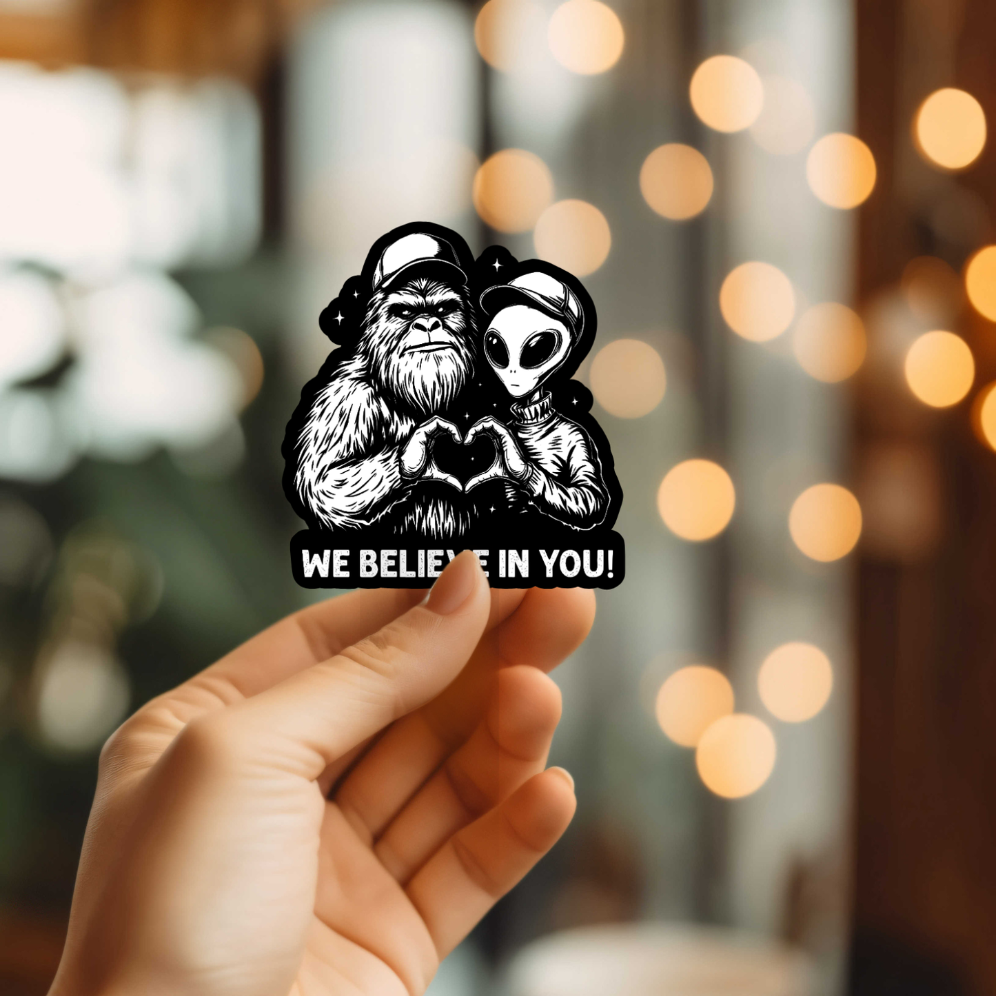 Hand holding a sticker with big foot and alien design and text 'WE BELIEVE IN YOU!' against a blurred background.