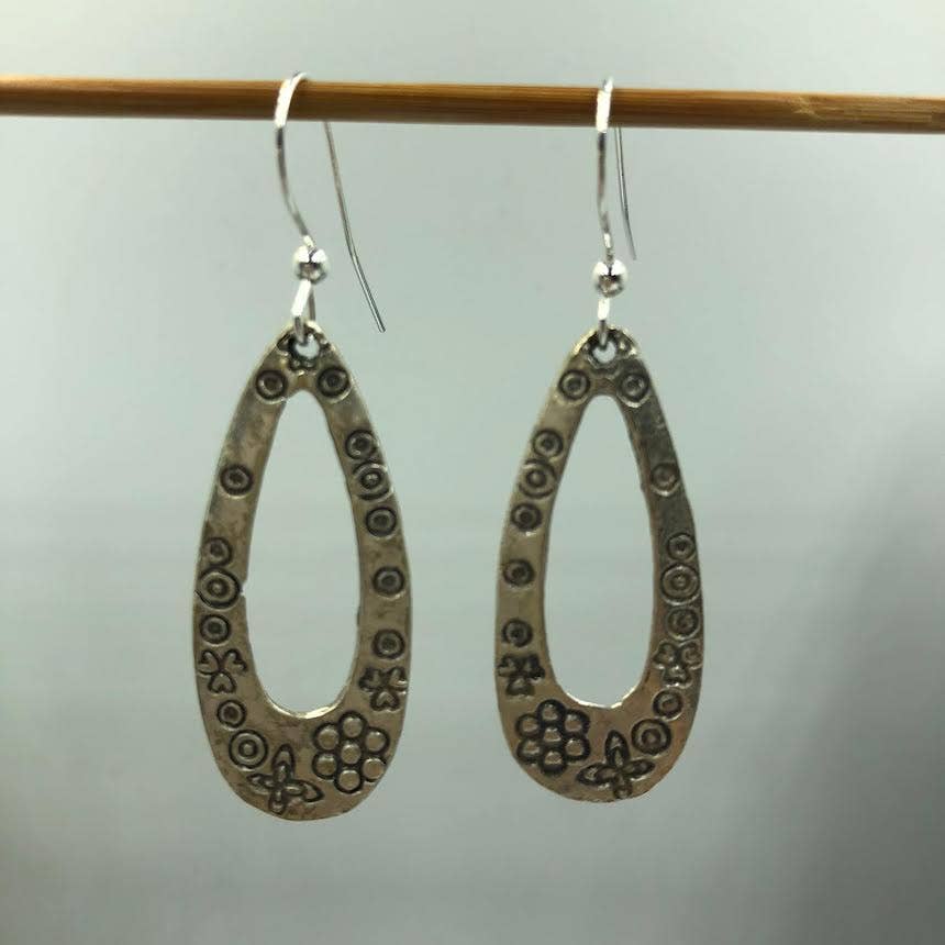 A pair of handmade teardrop-shaped dangle earrings made from oxidized silver metal, featuring stamped floral and circular patterns, hanging from a wooden rod against a neutral gray background.