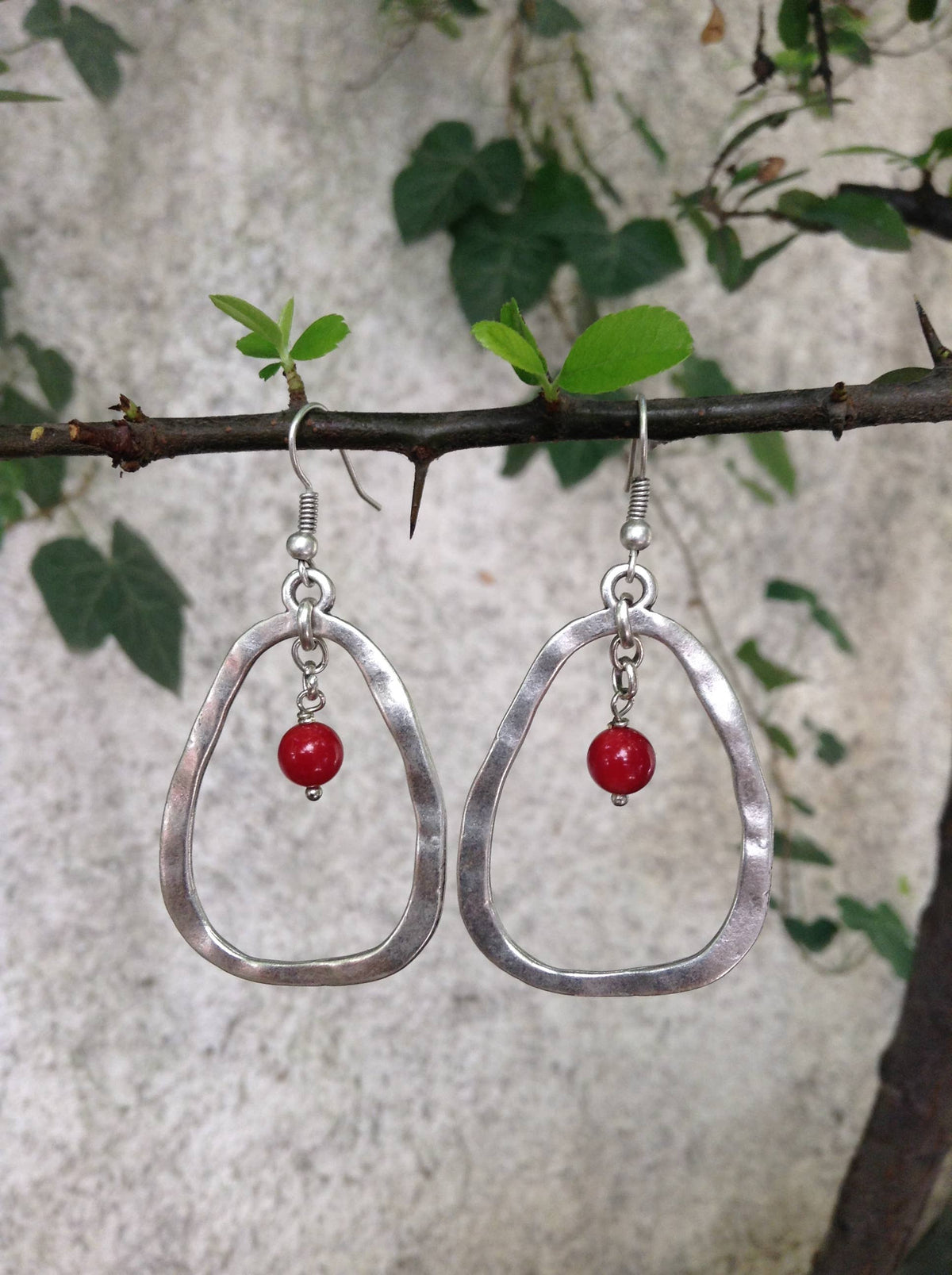 A pair of silver-tone hammered oval drop earrings with a small red bead dangle, hanging from a tree branch against a textured light gray wall with green leaves.
