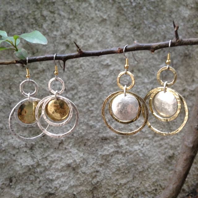 Pair of gold and silver circular earrings hanging from a branch against a stone wall.