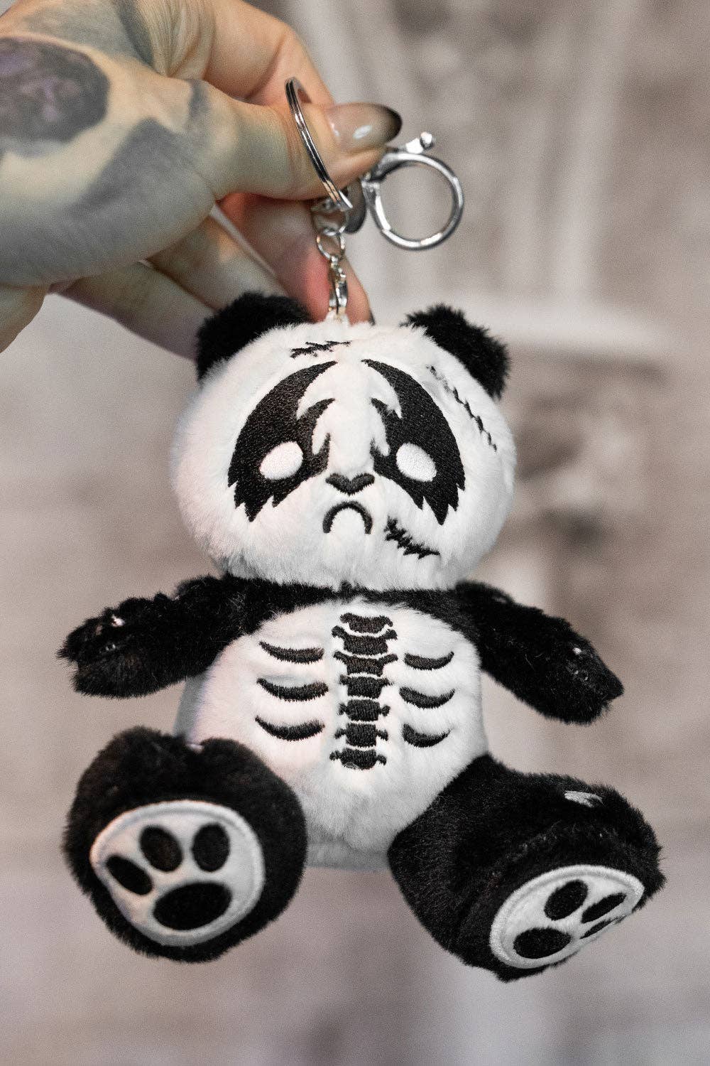 Plush keychain panda with skeleton design held by a hand against a blurred background