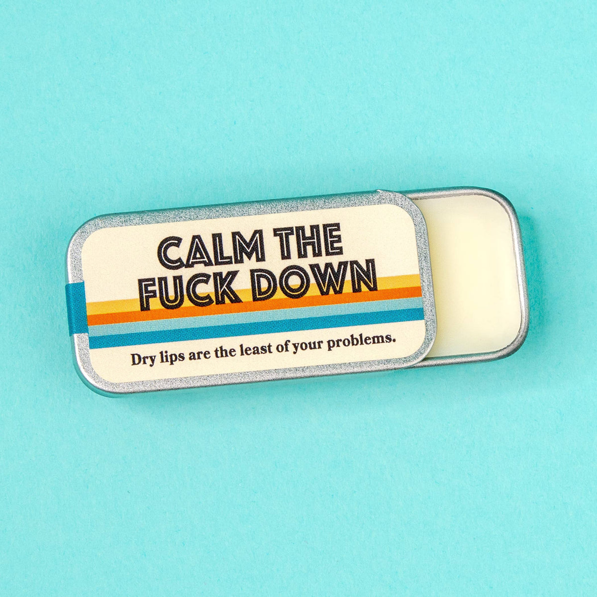 A small silver metal tin of lip balm with a cream-colored label that reads 'CALM THE FUCK DOWN' and 'Dry lips are the least of your problems.' The tin is shown open with the balm visible, set against a solid light blue background.