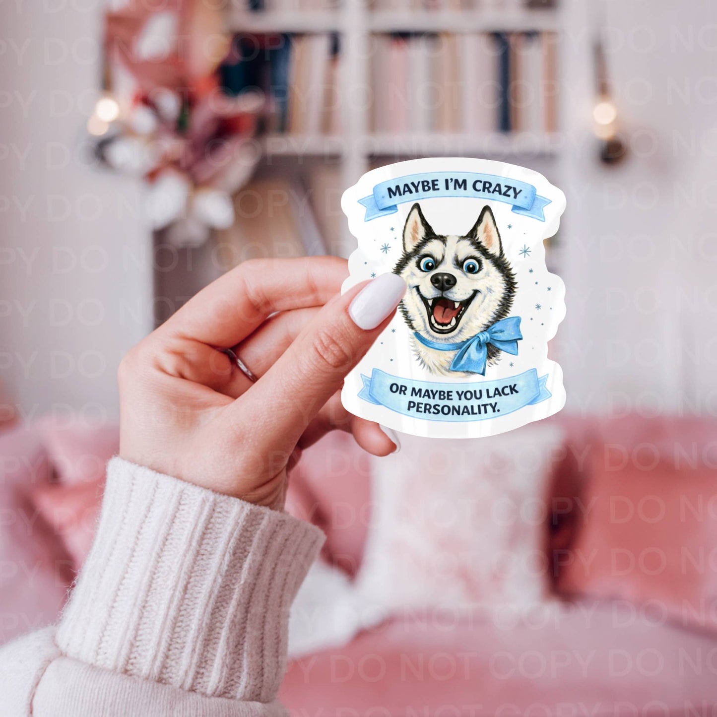 Hand holding a sticker with a Husky dog illustration and text "Maybe I'm Crazy, or maybe you lack personality" with a blurred indoor background.