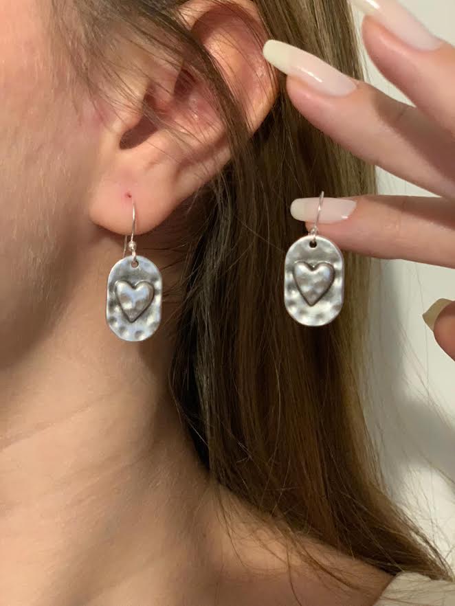 A close-up of a person wearing silver dangle earrings with a hammered oval shape and a raised heart design in the center, with a hand holding a matching earring beside the ear.