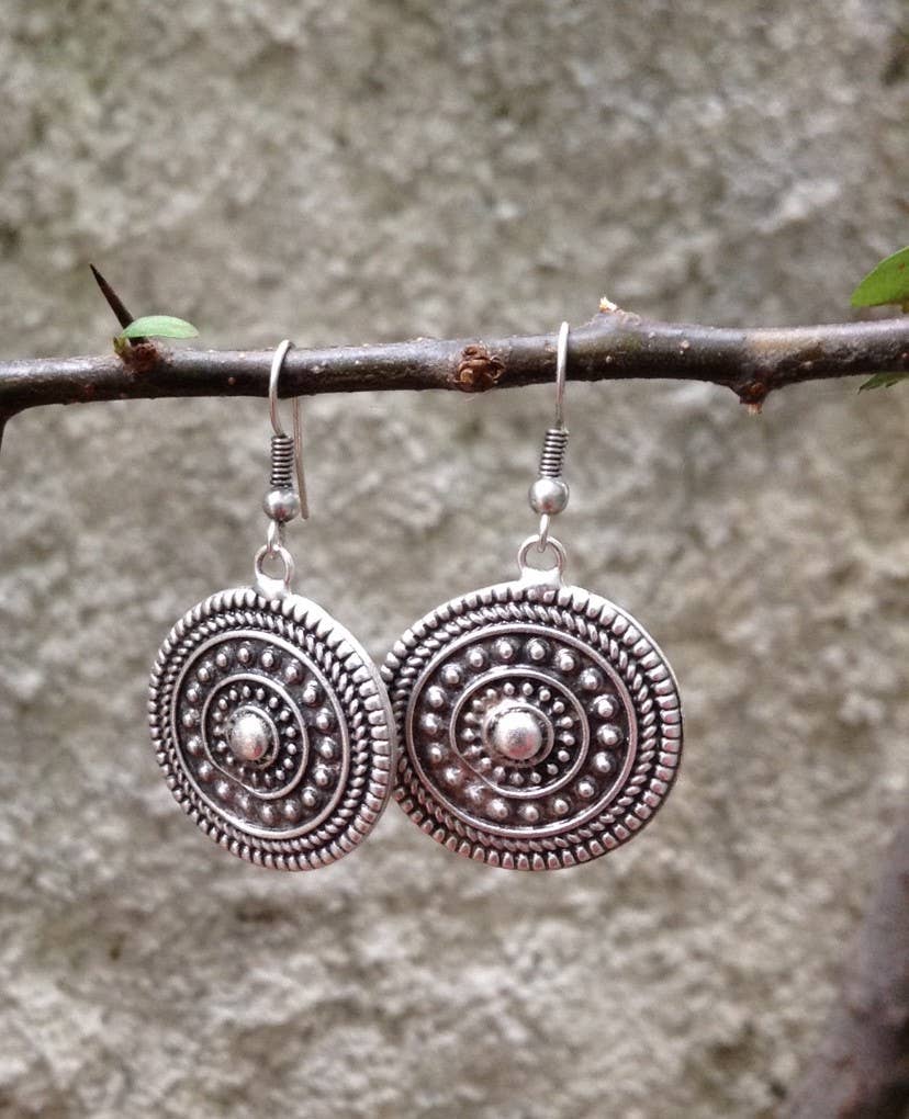 A pair of silver-toned circular drop earrings with intricate textured concentric patterns and small central beads, hanging from a natural twig against a blurred gray stone wall background.