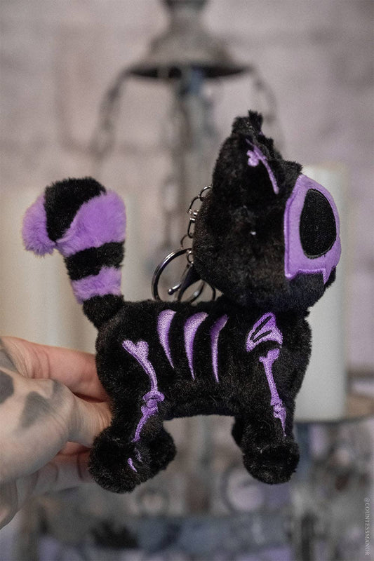 Black plush cat keychain with purple accents held by a hand against a blurred background