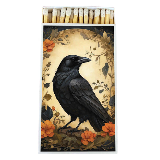 A matchbox with a printed illustration of a black raven surrounded by orange flowers and dark foliage, shown with a row of matches at the top on a white background.