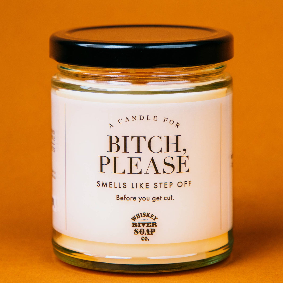 A jar candle with a black lid and a white label reading 'A CANDLE FOR BITCH, PLEASE SMELLS LIKE STEP OFF Before you get cut.' and 'Whiskey River Soap Co.
