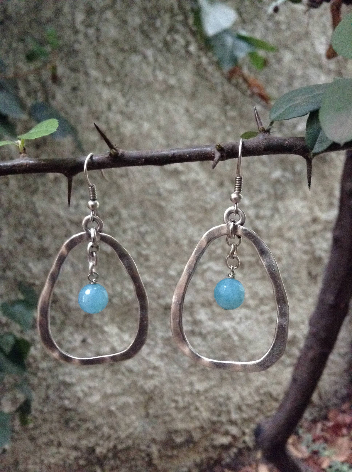 A pair of silver-toned teardrop-shaped dangle earrings with a small light blue bead, hanging from a natural branch with thorns against a blurred stone wall and green leaves.