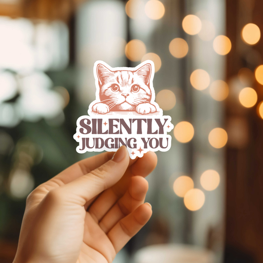 Hand holding a sticker with a cat illustration and text 'Silently Judging You' against a blurred background.