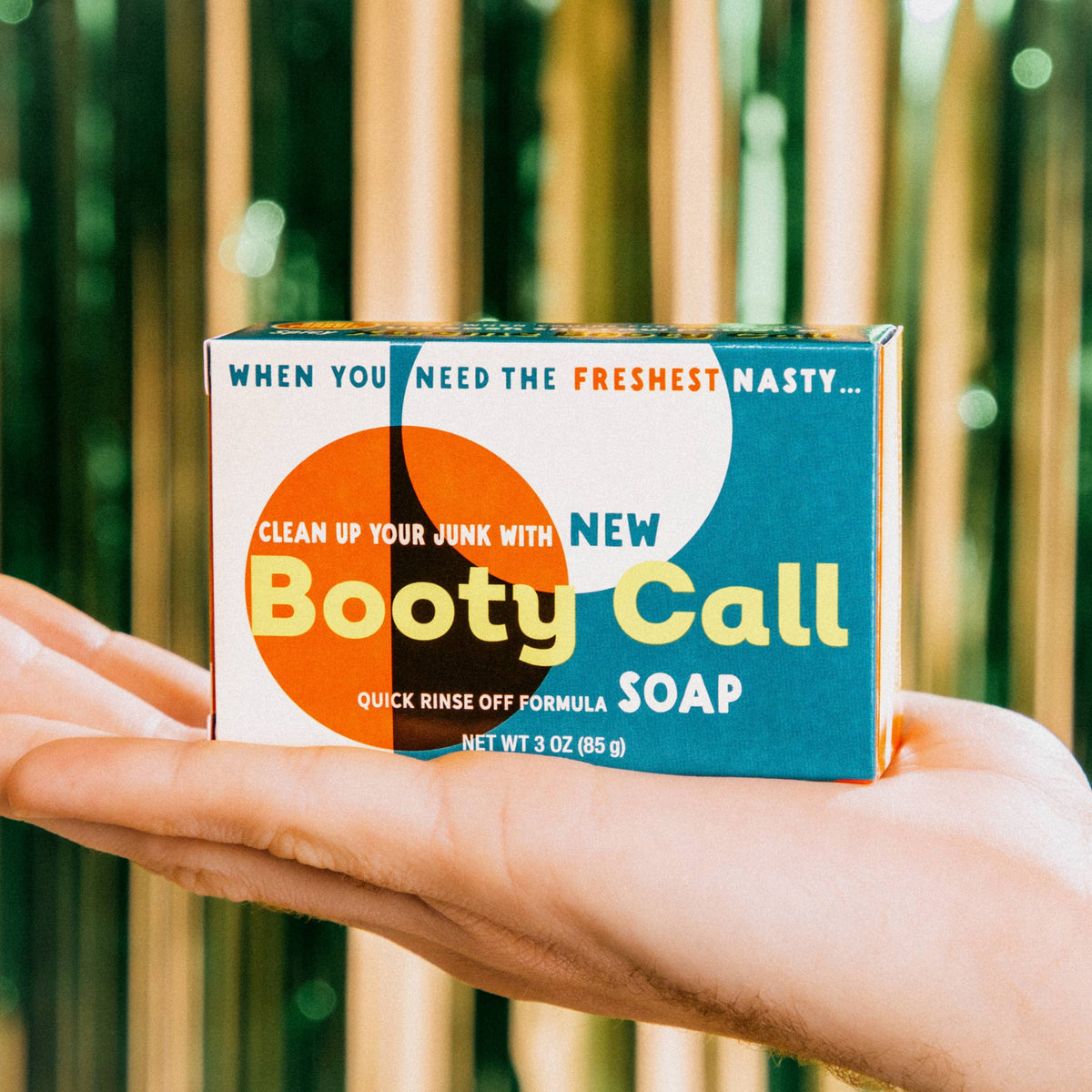 Hand holding a box of 'Booty Call' soap with a colorful label against a blurred natural background.