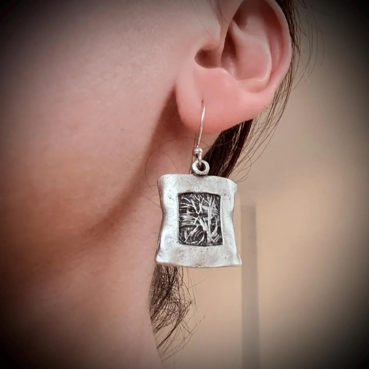 A close-up of a person's ear wearing a textured silver drop earring with a square pendant featuring an engraved tree design.