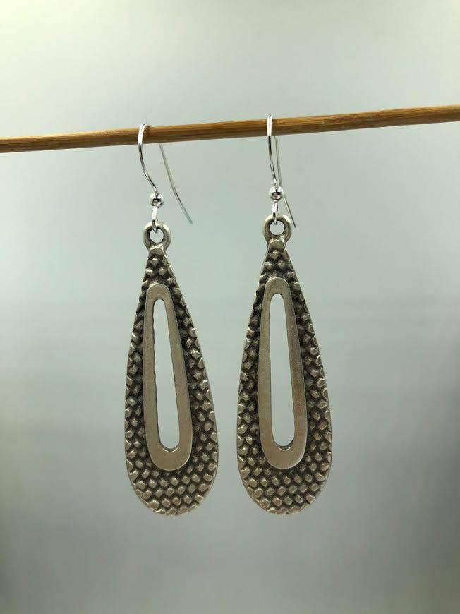 A pair of textured silver teardrop dangle earrings with a central cutout, hanging from a wooden rod against a light gray background.