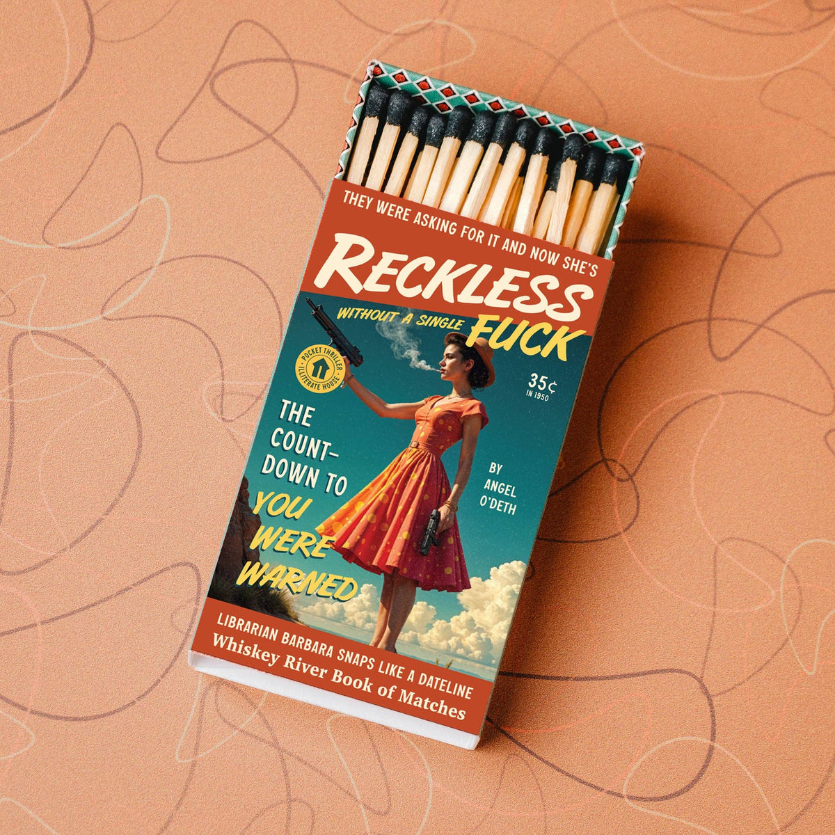 A novelty matchbox titled "Reckless Luck" lies open on a peach-colored surface with abstract line drawings. The matchbox cover features a retro illustration of a woman in a red dress smoking, with bold yellow and white text.