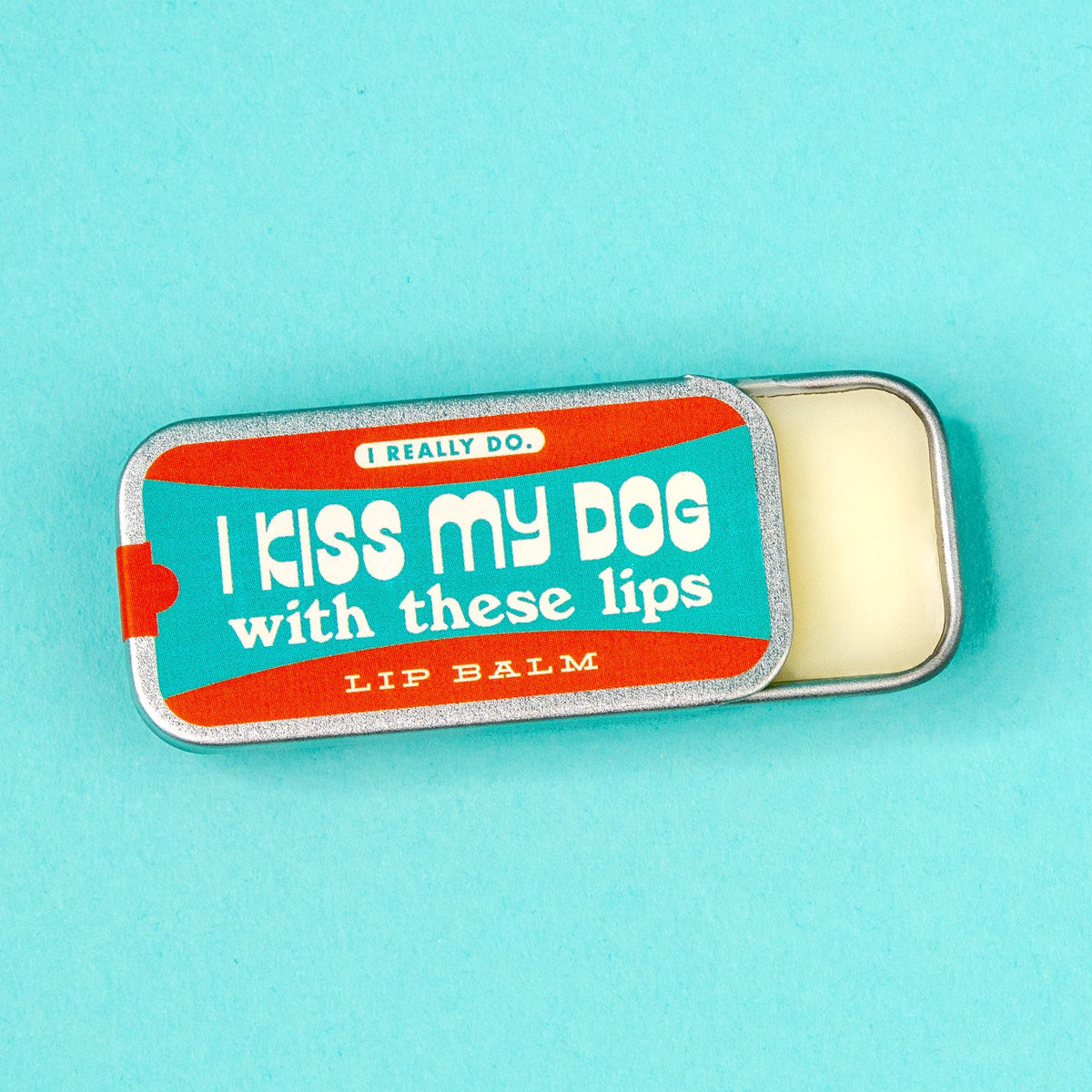 A tin of lip balm with the lid partially removed, showing the cream-colored balm inside. The lid features a colorful label with the text 'I kiss my dog with these lips."