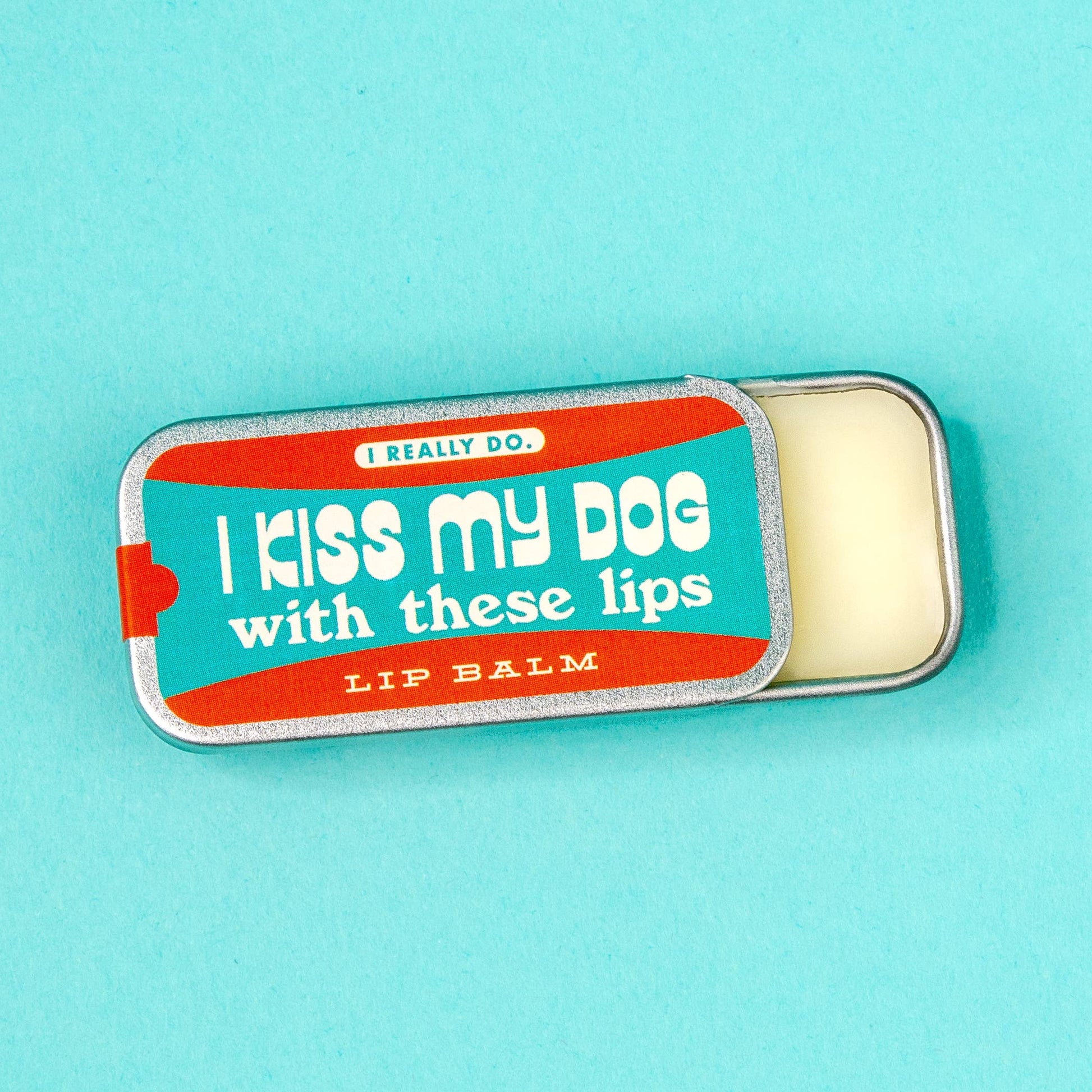 A tin of lip balm with the lid partially removed, showing the cream-colored balm inside. The lid features a colorful label with the text 'I kiss my dog with these lips."