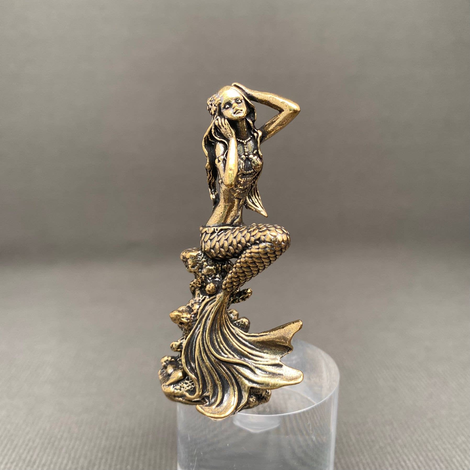 Brass mermaid figurine on a clear stand against a gray background