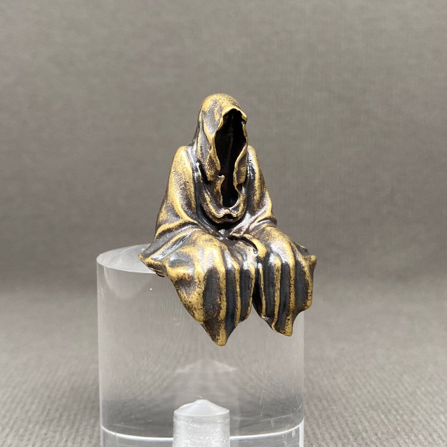 Brass statue of a hooded figure on a clear stand against a gray background