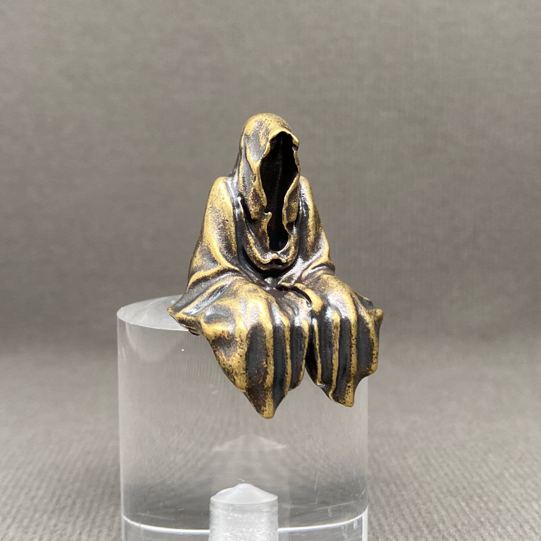 Brass statue of a hooded figure on a clear stand against a gray background