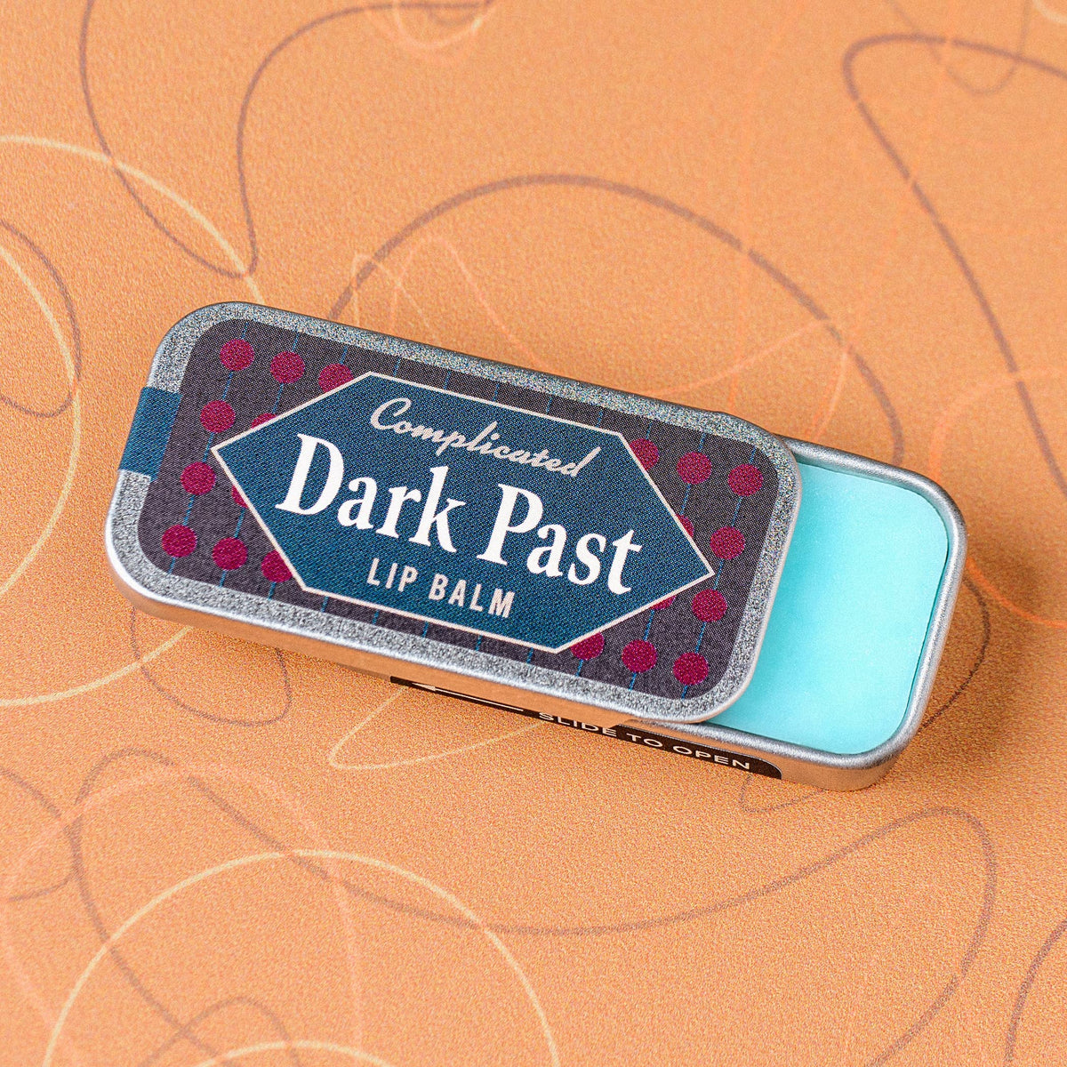 Lip balm tin labeled 'Dark Past' on an orange background