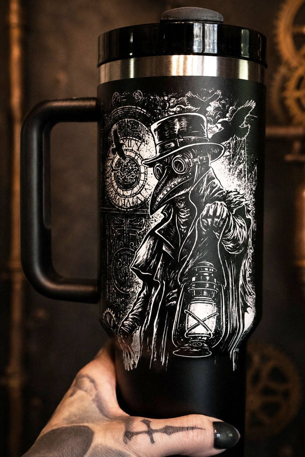 A black handled travel tumbler with a detailed white steampunk plague doctor illustration, held by a tattooed hand against a dark, vintage-themed background.