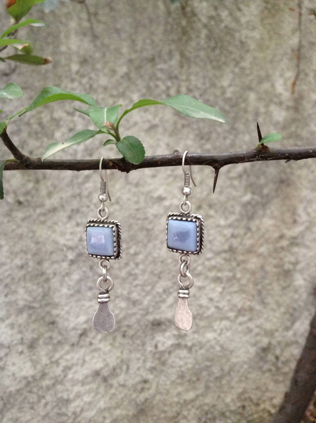 A pair of silver dangle earrings with rectangular light blue gemstones and teardrop-shaped silver charms, hanging from a tree branch against a textured gray stone wall.