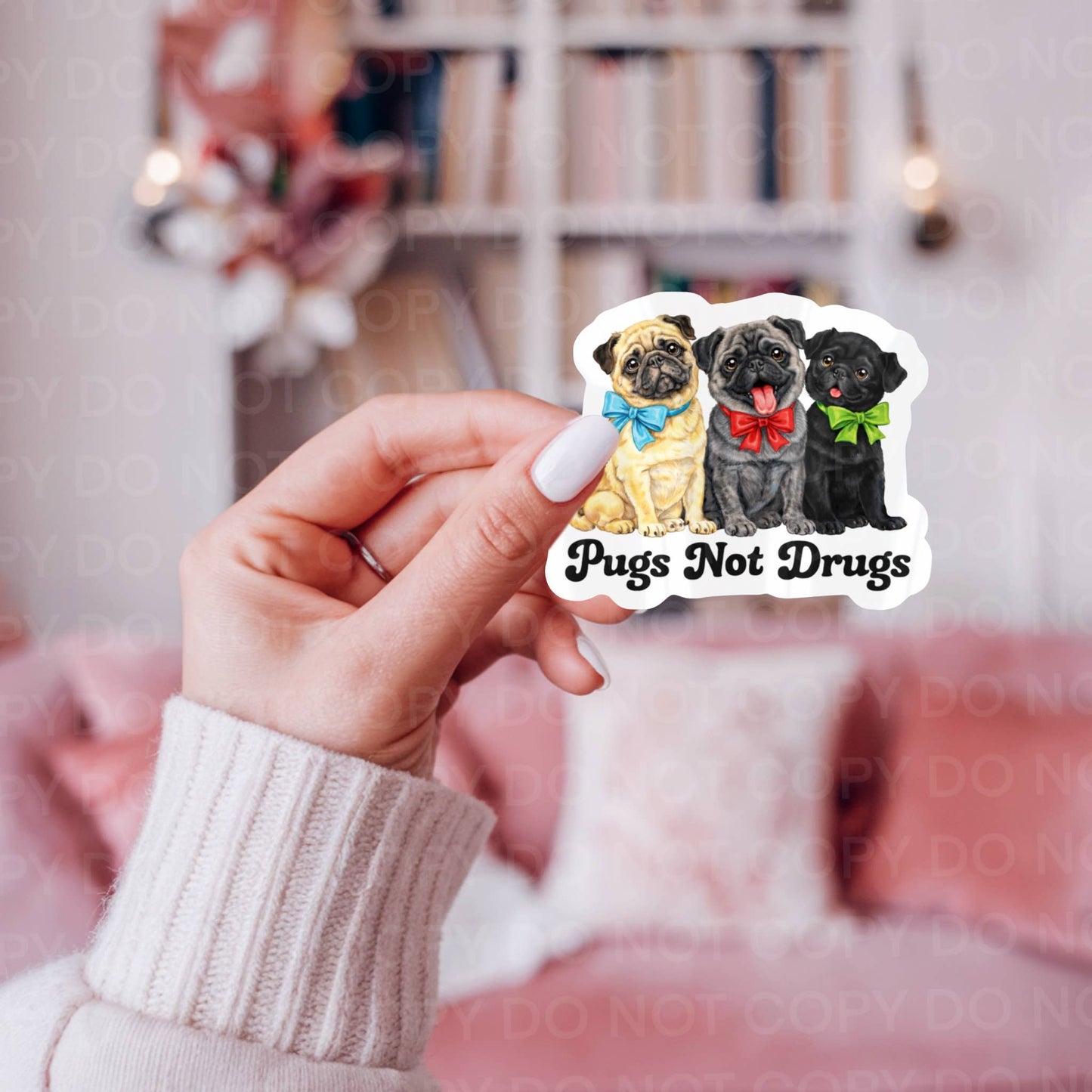 Hand holding a sticker with three pugs and 'Pugs Not Drugs' text against a blurred indoor background.