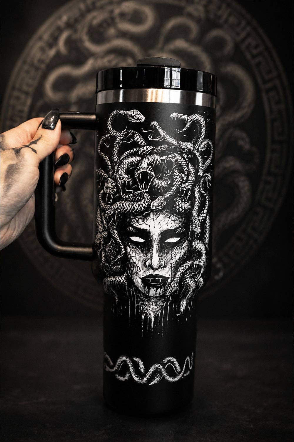 A black handled travel tumbler with a lid, featuring a detailed white gothic Medusa illustration, held by a hand with black fingernails against a dark background with a circular pattern.