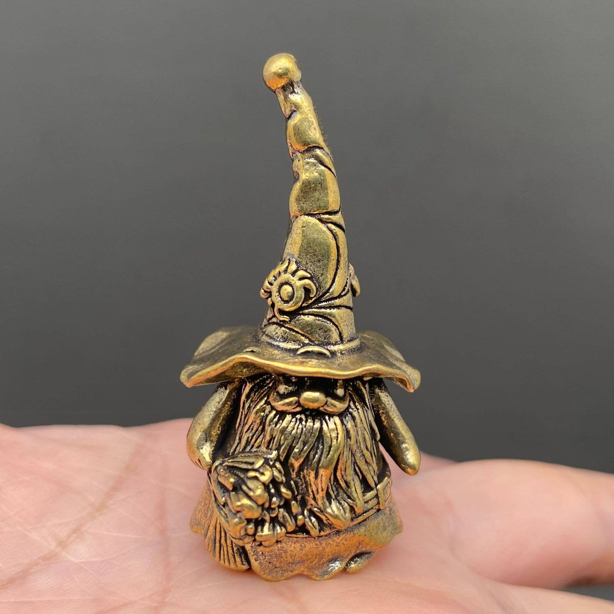 A small brass gnome figurine with a pointed hat and long beard, resting on a person's palm against a dark gray background.