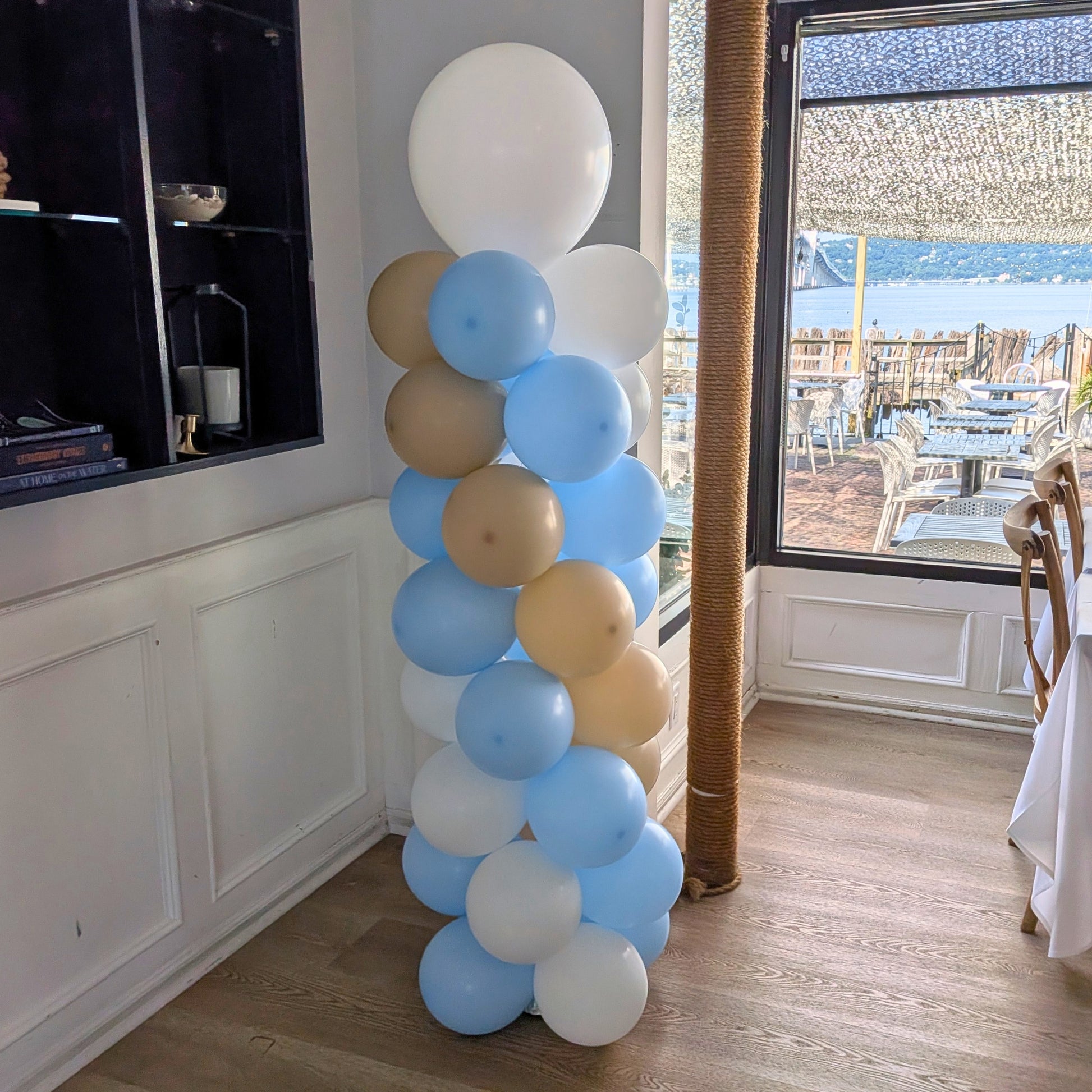 Photo of a balloon column made with blue, white, and tan latex balloons standing in a restaurant with wood floors.