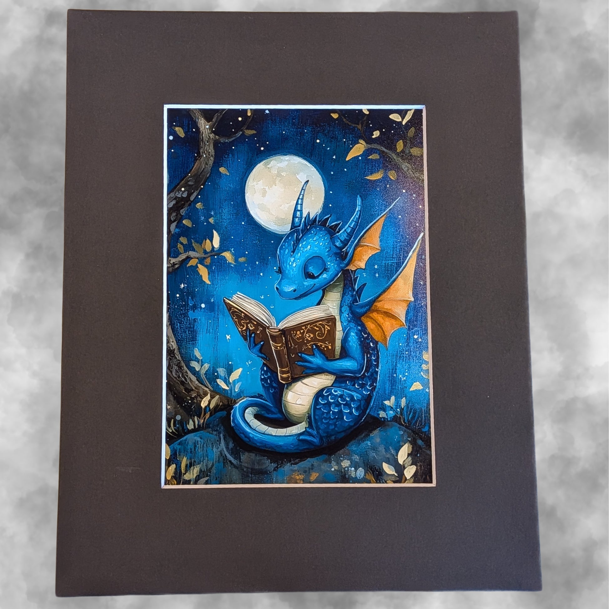 A square dark brown art print or card featuring a whimsical illustration of a blue dragon reading a book under a full moon, set against a mottled light gray background.