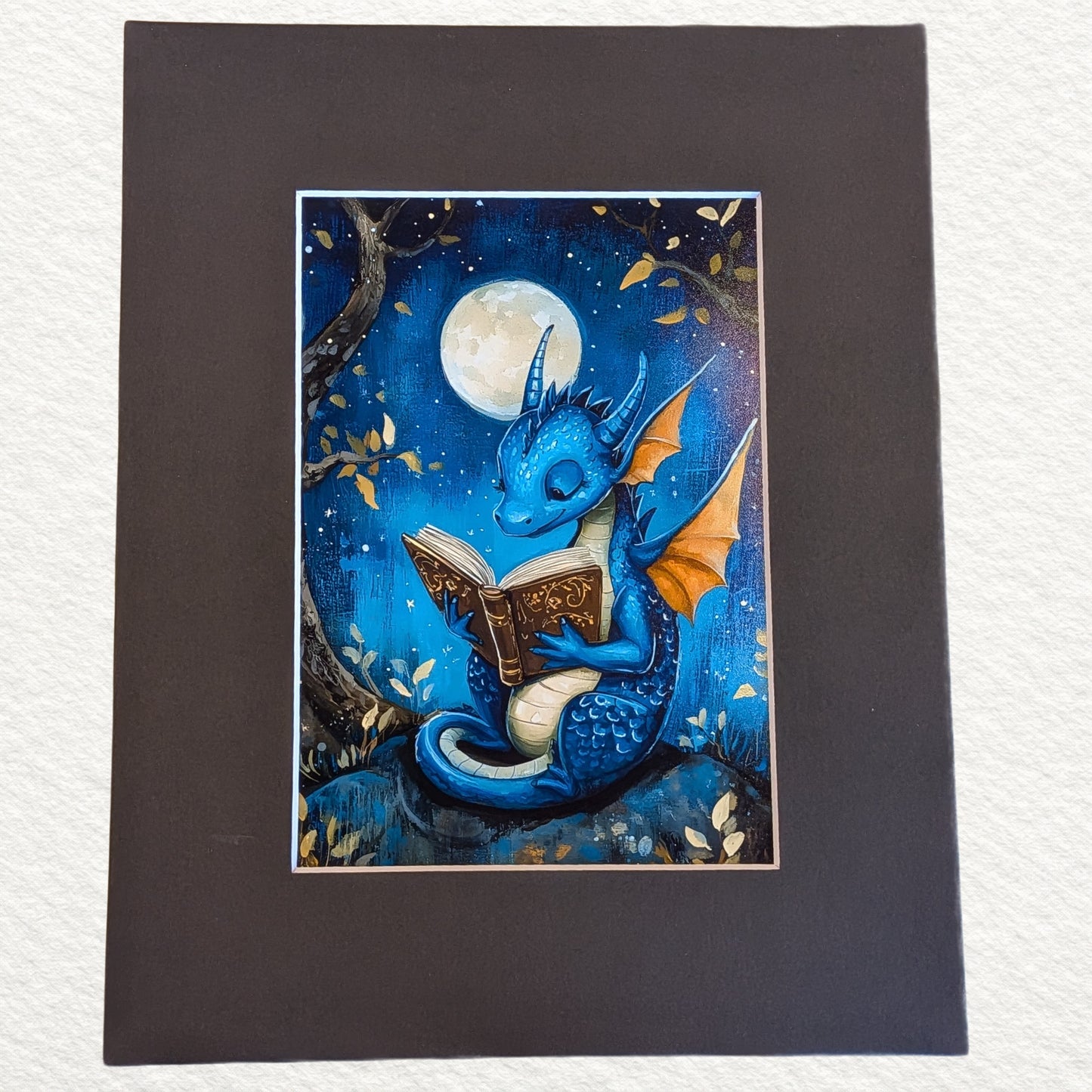 A square art print featuring a blue dragon reading a book under a full moon, mounted on a wide dark brown mat against a white textured background.