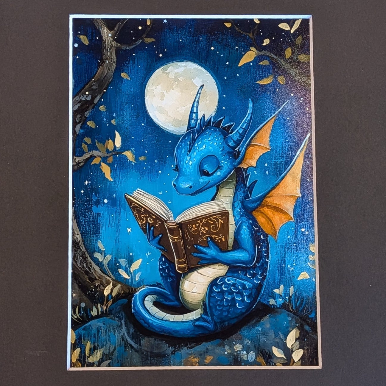 A matted art print featuring a whimsical illustration of a blue dragon reading a book under a full moon, set against a dark charcoal mat on a textured concrete floor.