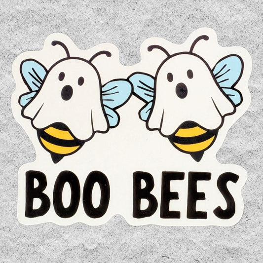 A die-cut sticker featuring two cartoon ghost bees with light blue wings and yellow-and-black striped bodies, above the words “BOO BEES” in large black letters on a white sticker border, shown against a light gray textured background.
